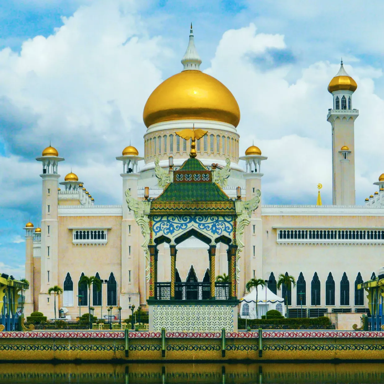 A majestic view of Sultan Omar Ali Saifuddien Mosque in Brunei, featuring its gleaming golden domes and white marble structure. The mosque stands on a reflective lagoon with a grand royal barge in front, framed by blue skies and white clouds - on our Brunei work permit page.