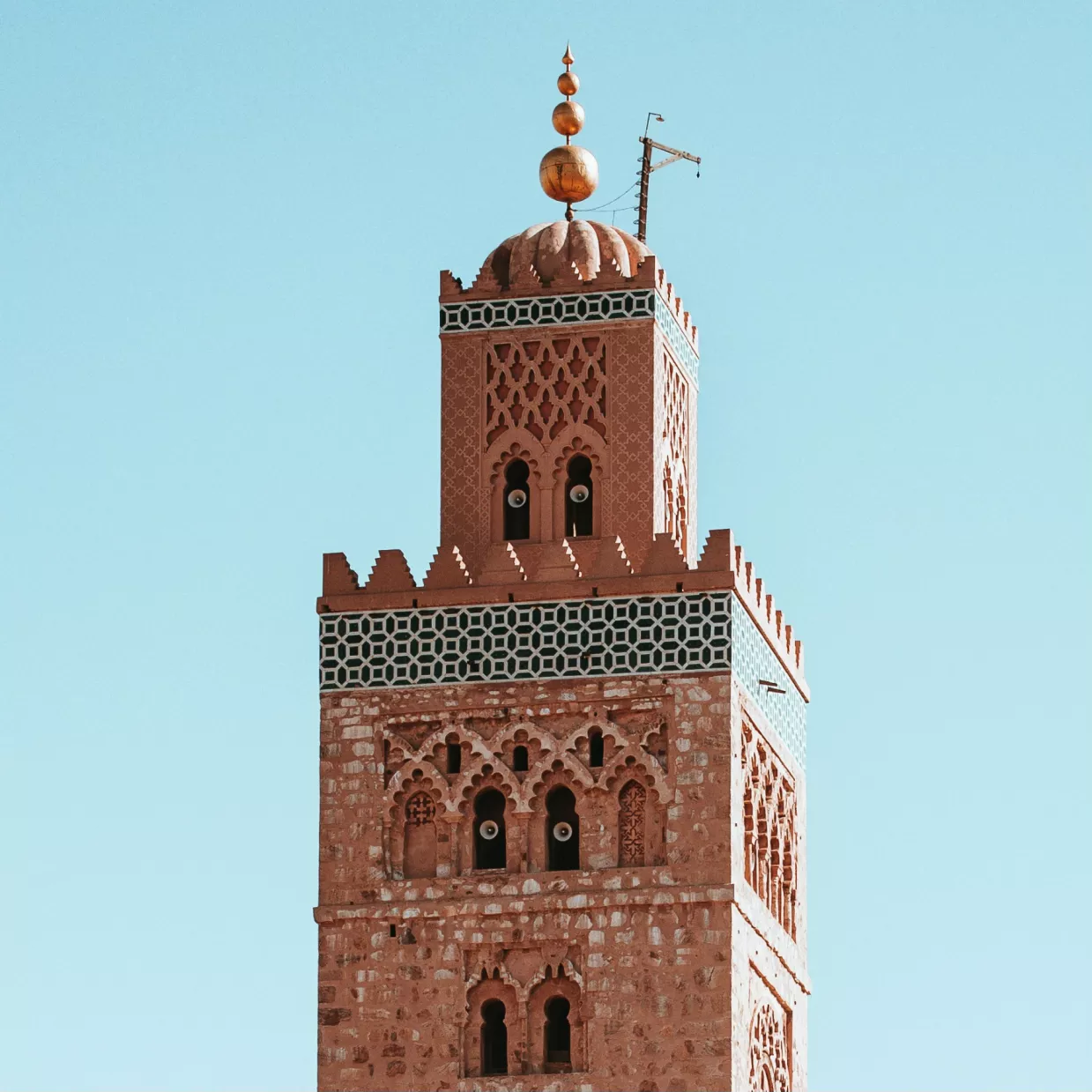 A detailed view of the Koutoubia Mosque in Marrakech, Morocco, showcasing its iconic red stone minaret adorned with intricate geometric designs. The minaret rises against a clear blue sky, crowned with copper globes, a symbol of Morocco's rich Islamic architecture and heritage.